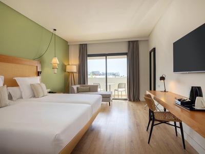 Helles Hotelzimmer mit Doppelbetten, Schreibtisch, TV und Balkon mit Aussicht.