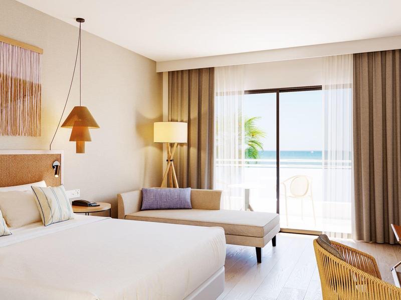 Helles Hotelzimmer mit großem Bett, Sofa, Stuhl, Schreibtisch und Balkon mit Meerblick.