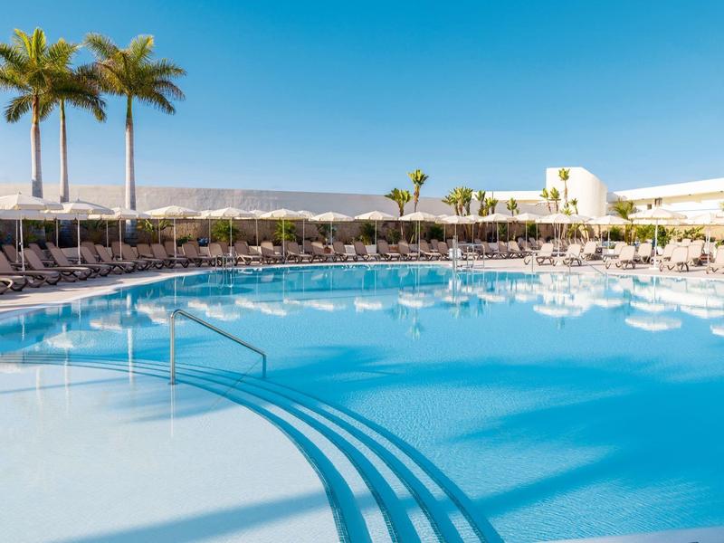 Großer Pool mit Sonnenschirmen und Palmen unter klarem blauem Himmel.