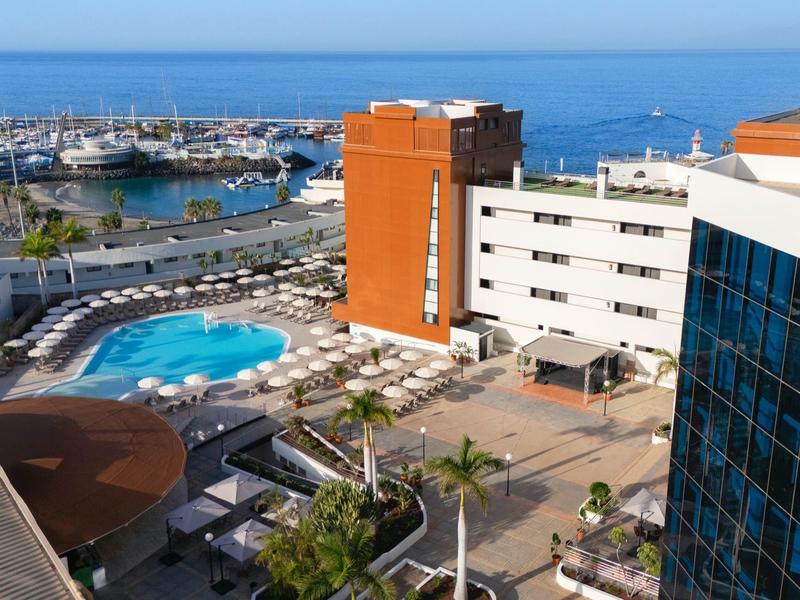 Hotel mit blauem Pool, weißen Liegestühlen, Palmen und Blick auf das blaue Meer im Hintergrund.