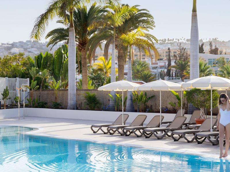 Ein Pool mit blauer Wasseroberfläche neben Sonnenliegen und Palmen bei Sonnenschein.