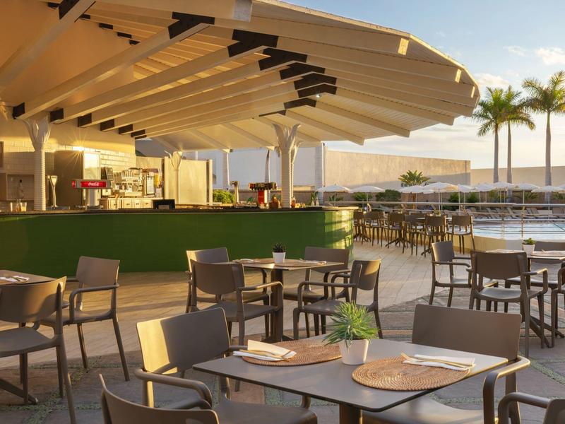 Moderner Außenbereich mit Tischen, Stühlen und Palmendekor neben einem Pool bei Sonnenuntergang.