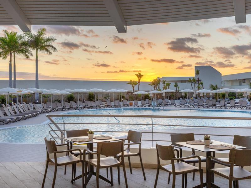 Moderner Außenpoolbereich mit Tischen, Stühlen und Palmen bei Sonnenuntergang.