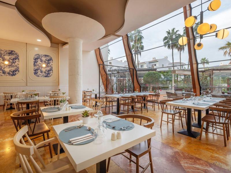 Helles Restaurant mit großen Fenstern, modernen Holzmöbeln und rundlichen Lampen an der Decke.