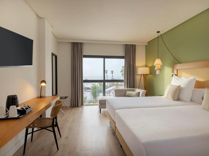 Modernes Hotelzimmer mit zwei Betten, Schreibtisch, TV und Balkon mit Blick auf das Wasser.