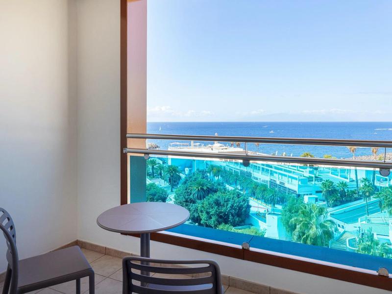 Balkon mit Tisch und Stühlen, Blick auf Poollandschaft und Meer unter klarem Himmel.