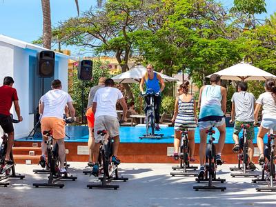 Skupina lidí cvičících venku na stacionárních kolech pod vedením instruktora, obklopena stromy.