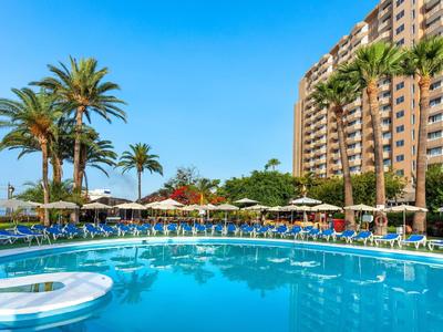 Hotel s velkým venkovním bazénem, lehátky a palmami pod jasnou modrou oblohou.