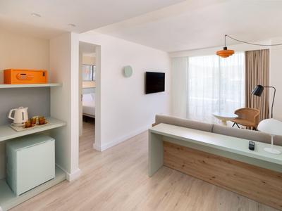 Heller Hotelzimmerbereich mit modernem Mobiliar, TV, Sessel und Balkon mit Tageslicht.