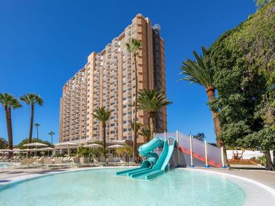Tall hotel building with pool, water slides, and palm trees under a clear blue sky.