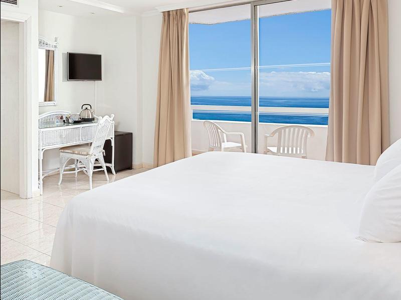 Helles Hotelzimmer mit großem Bett, Schreibtisch und Meerblick durch große Fenster.