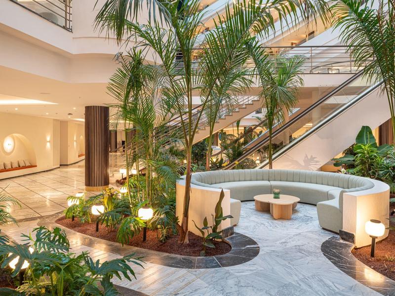 Hall d'hôtel moderne avec palmiers, zone d'assise incurvée et escalier en arrière-plan.