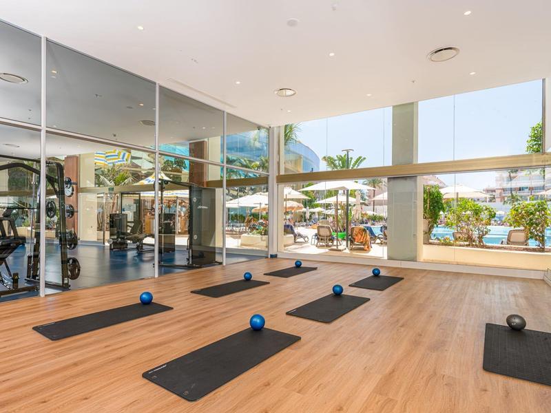 Salle de sport moderne avec grandes fenêtres, sol en bois, tapis de yoga et petits ballons.