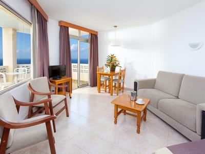 Helles Wohnzimmer mit grauem Sofa, Holztisch, Stühlen und Balkon mit Meerblick.