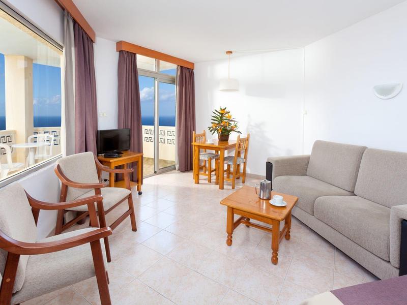 Helles Wohnzimmer mit grauem Sofa, Holztisch und zwei Stühlen, großem Fenster mit Blick auf das Meer.