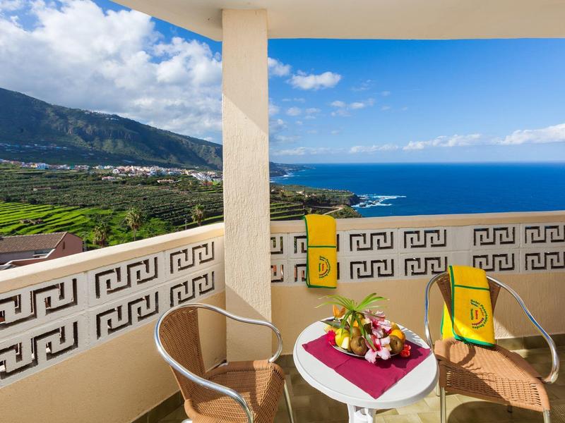 Balkon mit zwei Stühlen, Tisch, Blumen und Meerblick vor Bergen unter blauem Himmel.