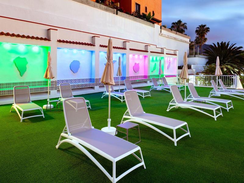 Mehrere weiße Liegestühle mit grauen Polstern stehen auf grünem Kunstgras vor einer bunt beleuchteten Gebäudewand.