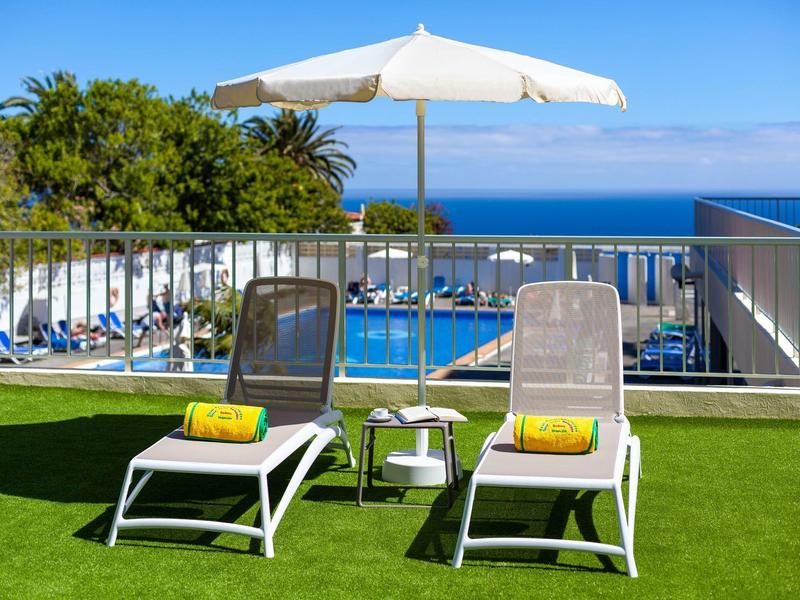 Zwei Liegestühle mit gelben Handtüchern unter einem weißen Sonnenschirm auf grünem Rasen, Meer im Hintergrund.