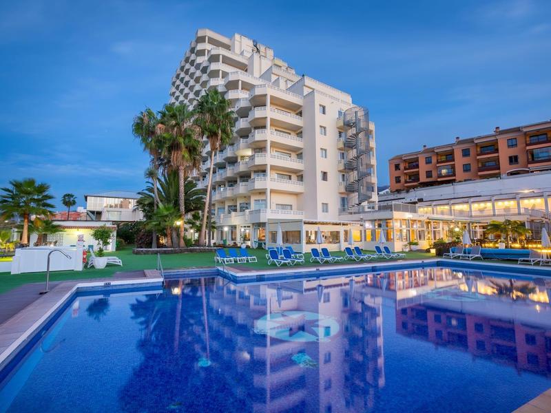 Moderner Apartmentkomplex mit großem Pool, Liegestühlen und Palmen unter blauem Himmel.