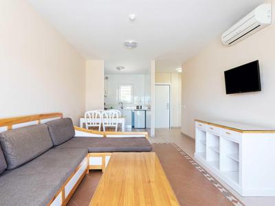 Salon lumineux avec canapé gris, table en bois, TV murale et coin repas en arrière-plan.