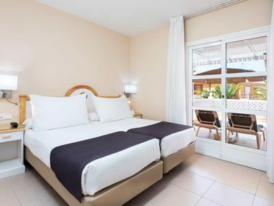 Chambre d'hôtel lumineuse avec lit double, murs blancs et accès à une terrasse avec chaises.