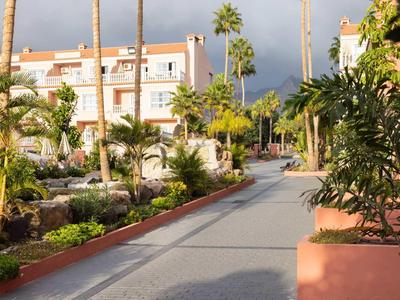 Large allée bordée de palmiers et végétation tropicale près d'un immeuble hôtelier sous un ciel nuageux.
