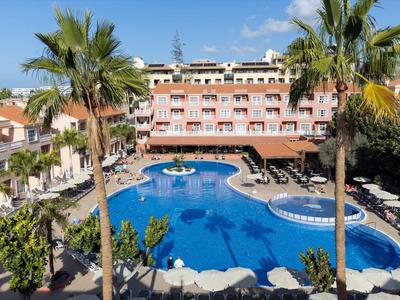 Hôtel avec grande piscine, palmiers et terrasse ensoleillée sous un ciel clair.