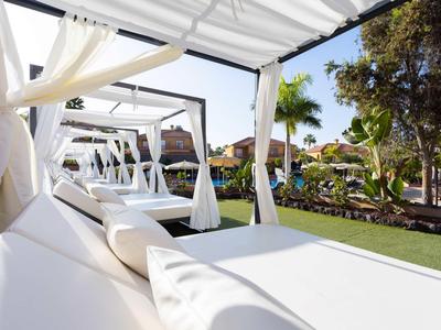 Lits lounge blancs avec des rideaux sous une pergola dans un jardin d'hôtel ensoleillé.