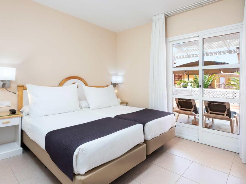 Chambre d'hôtel lumineuse avec lit double, murs blancs et accès à une terrasse avec chaises.