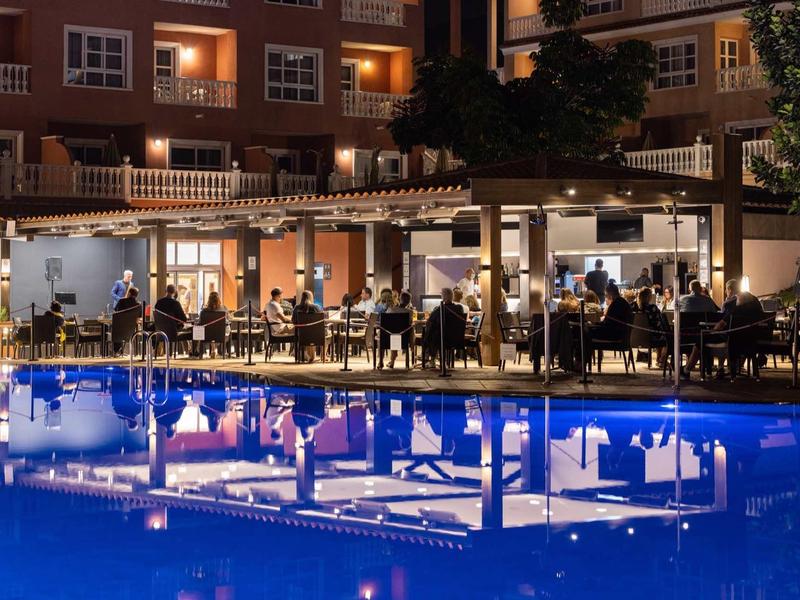 Terrasse d'hôtel avec des clients à côté d'une piscine éclairée la nuit.