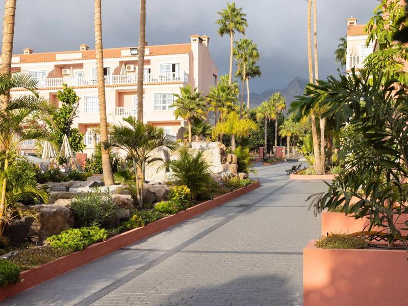 Large allée bordée de palmiers et végétation tropicale près d'un immeuble hôtelier sous un ciel nuageux.