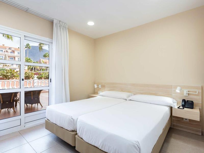 Chambre d'hôtel lumineuse avec deux lits et accès à un balcon avec chaises.