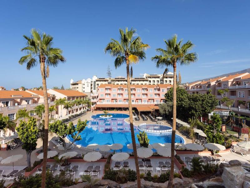 Grand hôtel avec piscine bleue entourée de palmiers et de chaises longues sous un ciel clair.