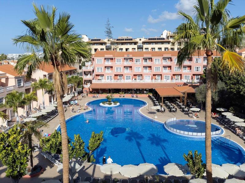 Hôtel avec grande piscine, palmiers et terrasse ensoleillée sous un ciel clair.