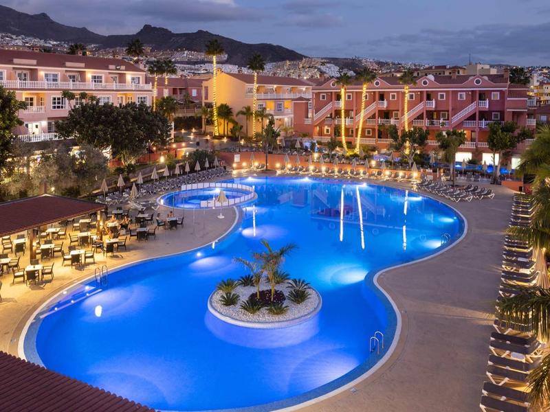 Grande piscine éclairée avec chaises longues dans un complexe hôtelier au crépuscule.