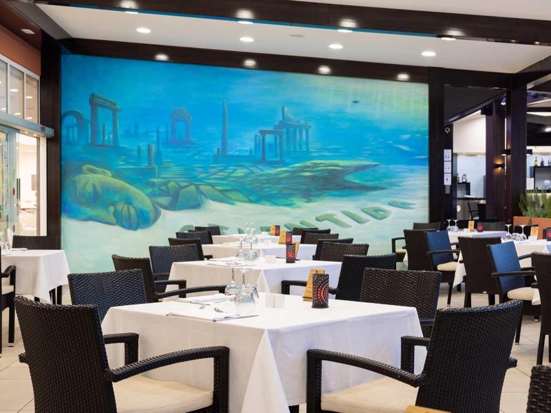 Restaurant avec des tables blanches dressées et une fresque murale de paysage côtier.