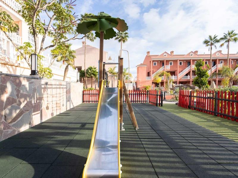 Aire de jeux pour enfants avec toboggan, sol en caoutchouc et palmiers en arrière-plan sous le soleil.