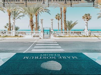 Een hotelentree met uitzicht op de zee, palmbomen en blauwe lucht.