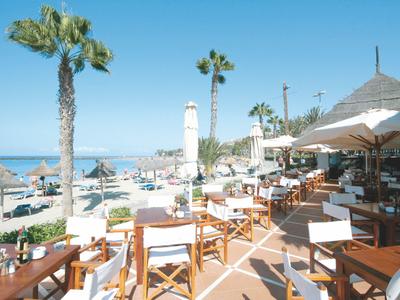 Strandrestaurant mit Holzstühlen, Sonnenschirmen, Palmen und Blick auf das Meer bei klarem Himmel.