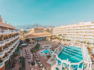 Großes Hotelgelände mit zwei Pools, vielen Liegestühlen und Bergen im Hintergrund unter blauem Himmel.