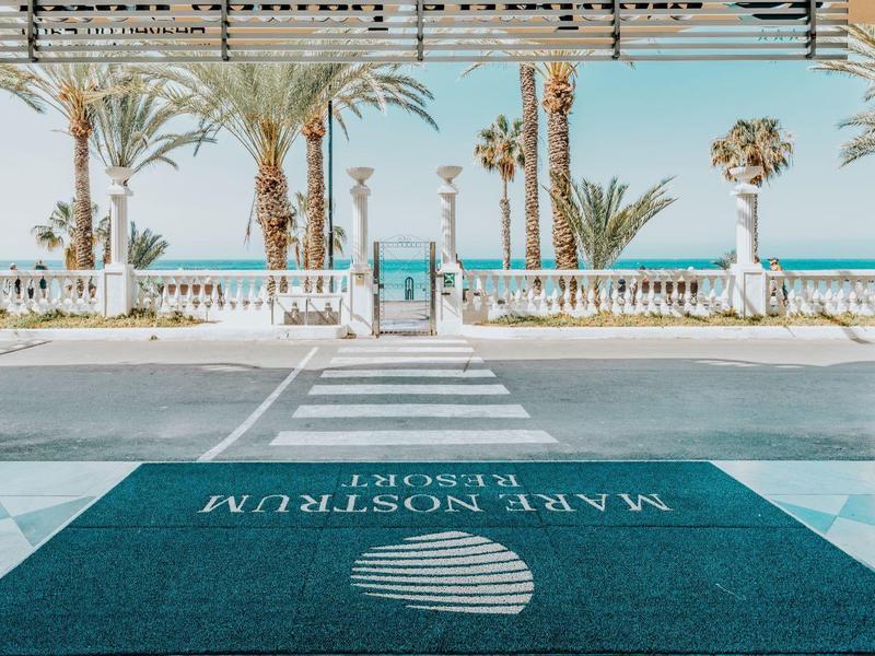 Heller, offener Eingangsbereich mit blauen Teppichen, weißen Säulen und Palmen vor strahlend blauem Himmel.