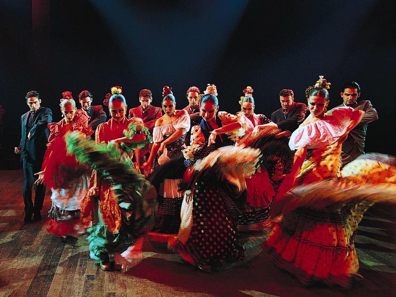 Bunte Flamenco-Tänzer in Tradition, lebhafte Bewegung, Holzfußboden und dunkler Theatersaal.