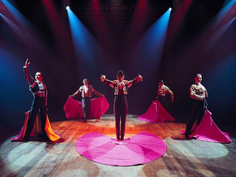 Fünf Flamenco-Tänzer auf Bühne mit roten Kleidern und dramatischem blauen Licht.