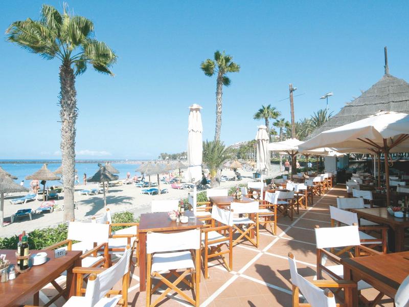 Strandrestaurant mit Holzstühlen, Sonnenschirmen, Palmen und Blick auf das Meer bei klarem Himmel.
