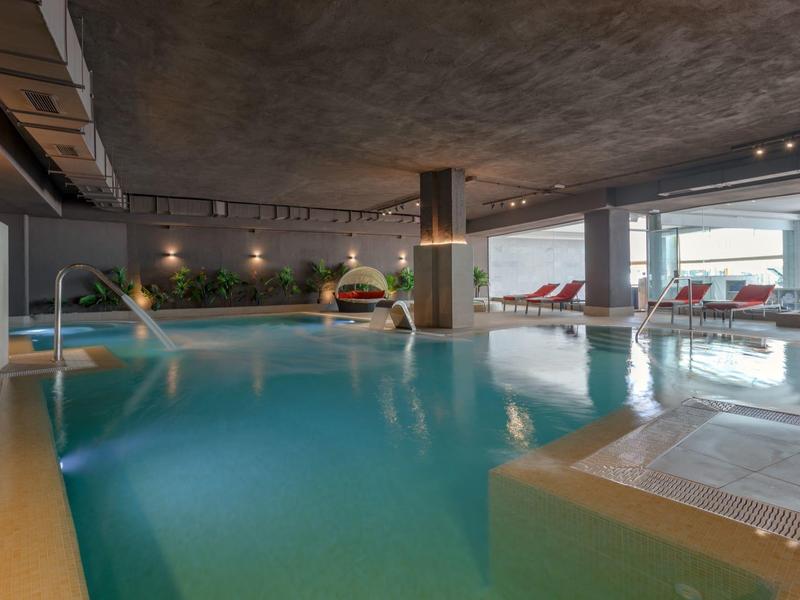 Innen-Schwimmbad mit großem Becken, Liegestühlen und Blick auf eine Terrasse mit Meer.