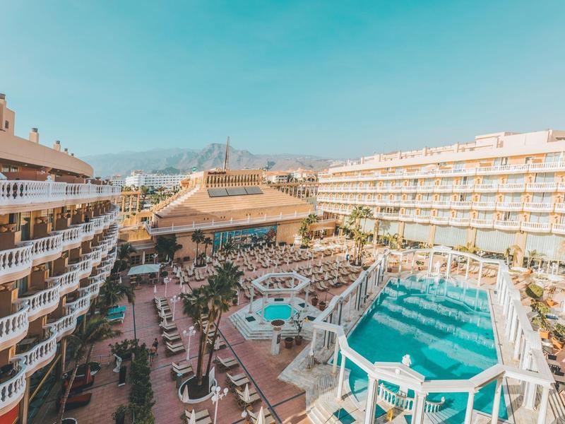 Großes Hotelgelände mit zwei Pools, vielen Liegestühlen und Bergen im Hintergrund unter blauem Himmel.