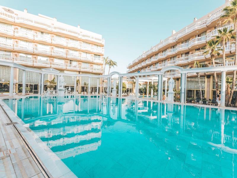 Großer blauer Pool zwischen zwei mehrstöckigen weißen Hotels unter klarem Himmel.