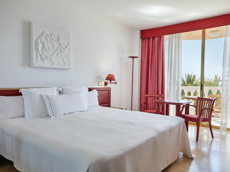 Hell eingerichtetes Hotelzimmer mit großem Bett, roten Möbeln und Balkon mit Meerblick.