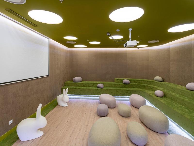 Moderner Raum mit grüner Wiese, weißem Bildschirm, runden Sitzkissen und Deckenspots.