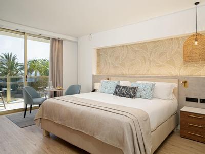 Moderne slaapkamer met groot bed, nachtkastje, stoelen en balkon met uitzicht op palmbomen.